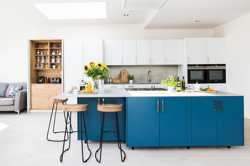 A bright kitchen with white cabinets on top and a blue kitchen island, Seats are arranged around the corner of one side of the island. 
