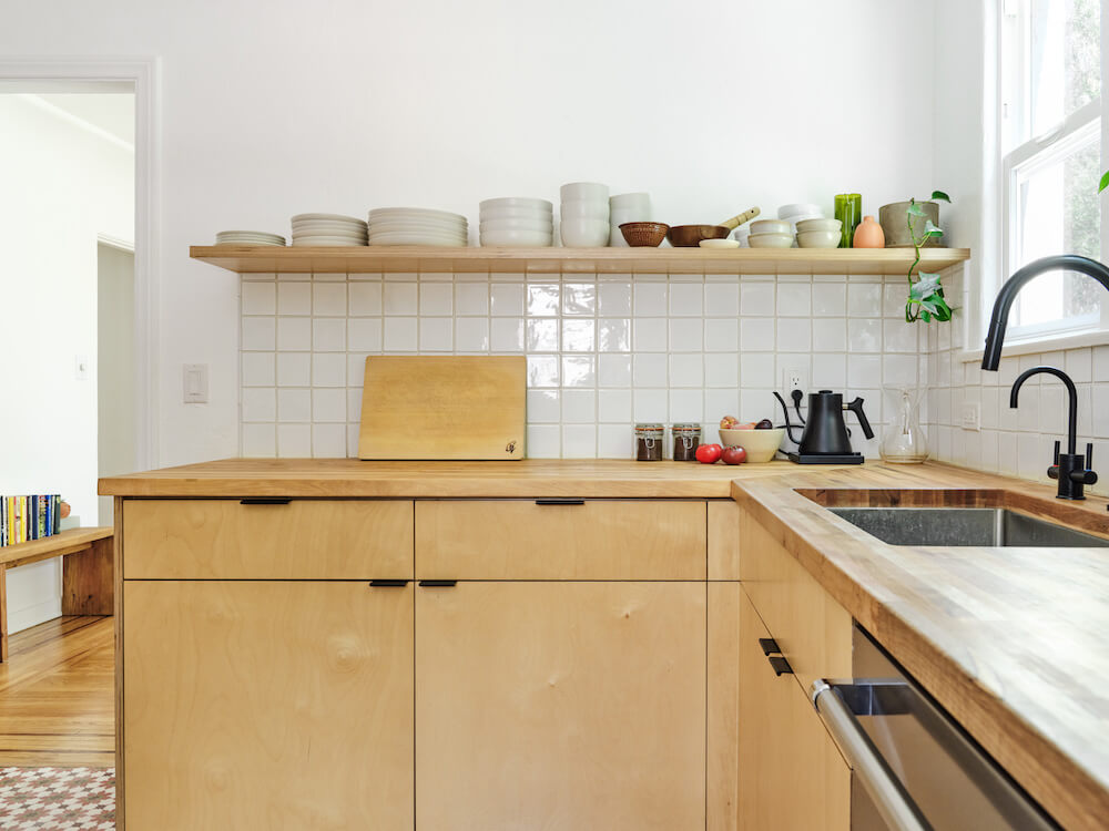 Open shelving holding bowls and plates over plywood kitchen cabinets and clay tile wall