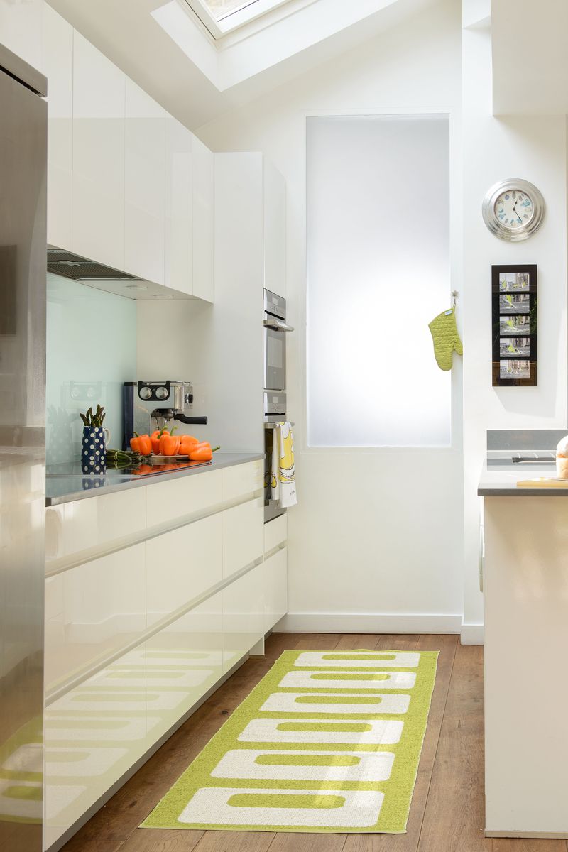 A minimalist galley kitchen with white cabinets and a bright green rug. 