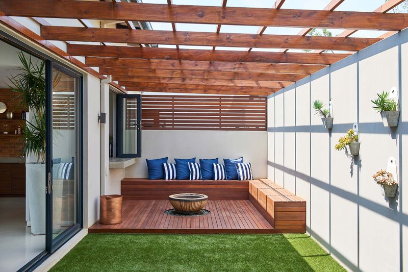 A wooden pergola attached to a house with a fire pit underneath and bench seating.