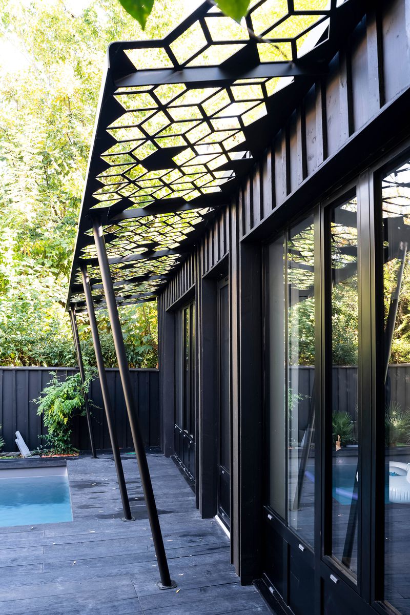 A modern metal pergola with a geometric cut out is attached to the side of a house, next to a pool. 