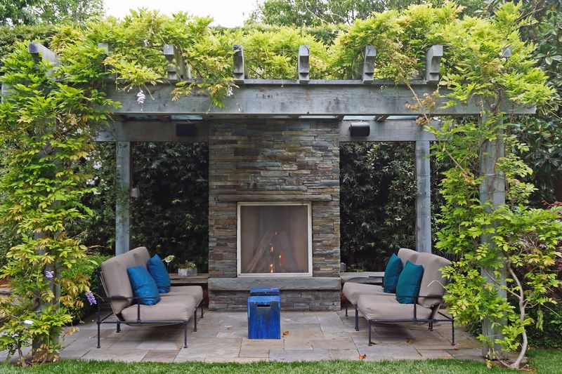 A wooden pergola with vines growing along the sides and top. Under the shaded area is stone fire place and seating.
