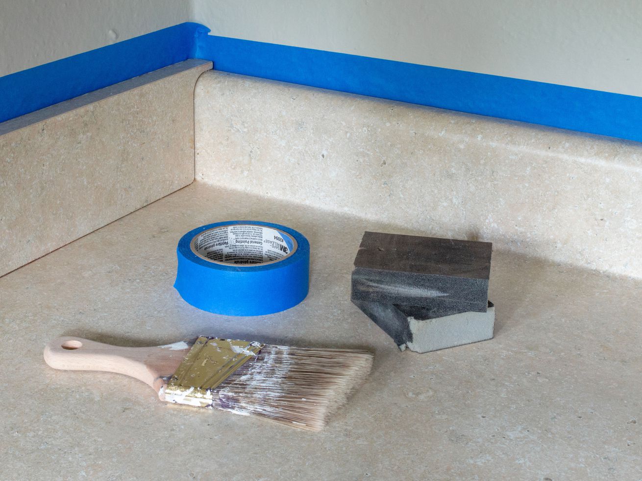 A paintbrush, blue tape, and sanding blocks sit on a beige laminate countertop. 