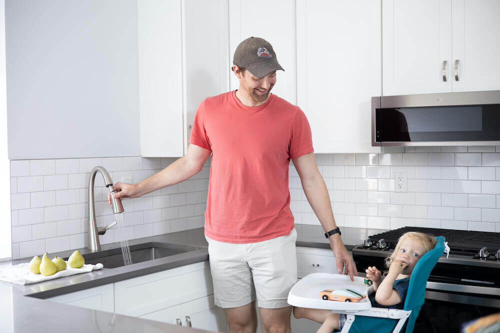 Image of a father feeding a baby in a high chair by a white kitchen sink