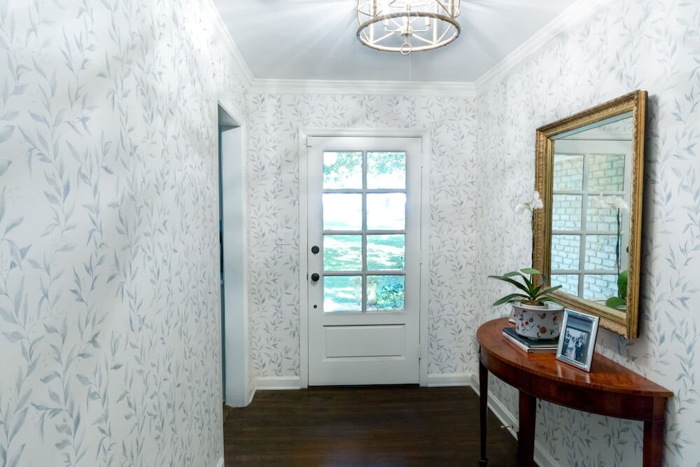 The entryway with leafy wallpaper, mirror, and table with a plant