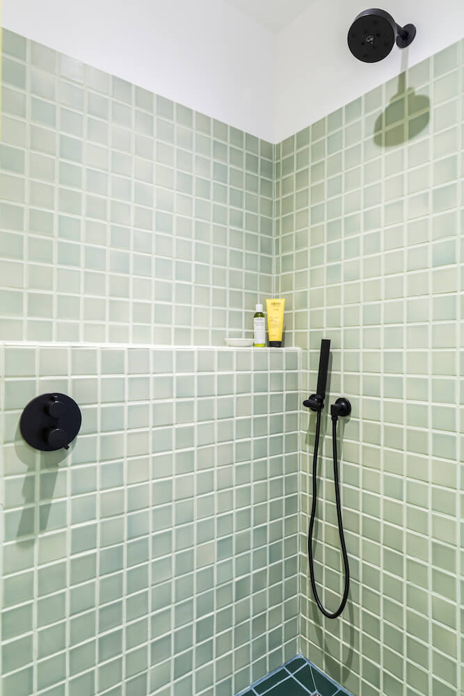 Image of a shower with light green tiles, black fixtures and shelf for bathroom supplies